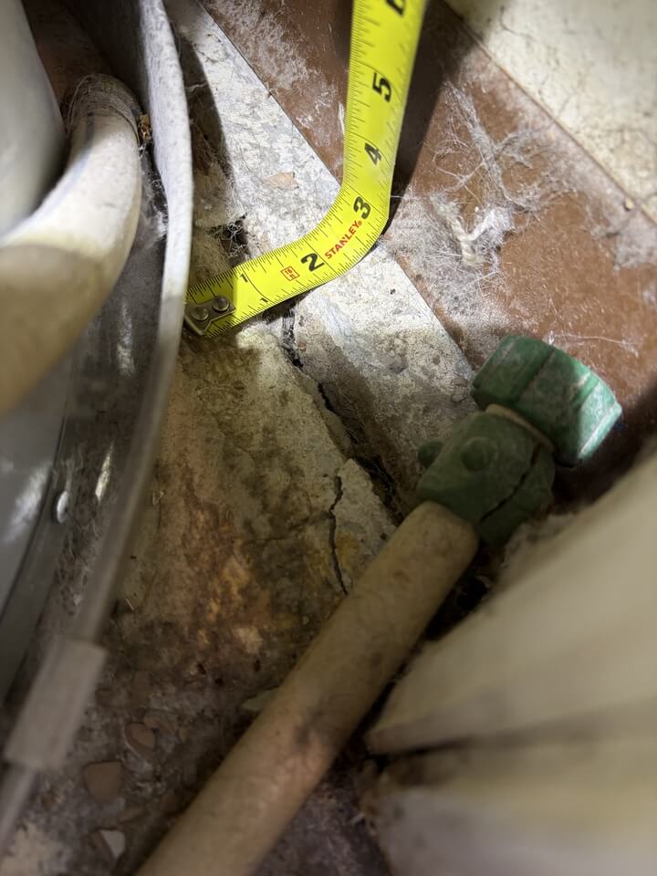 TTape measure beside one of the cracks in the garage floor, held against a gap in the concrete next to a pipe and green valve